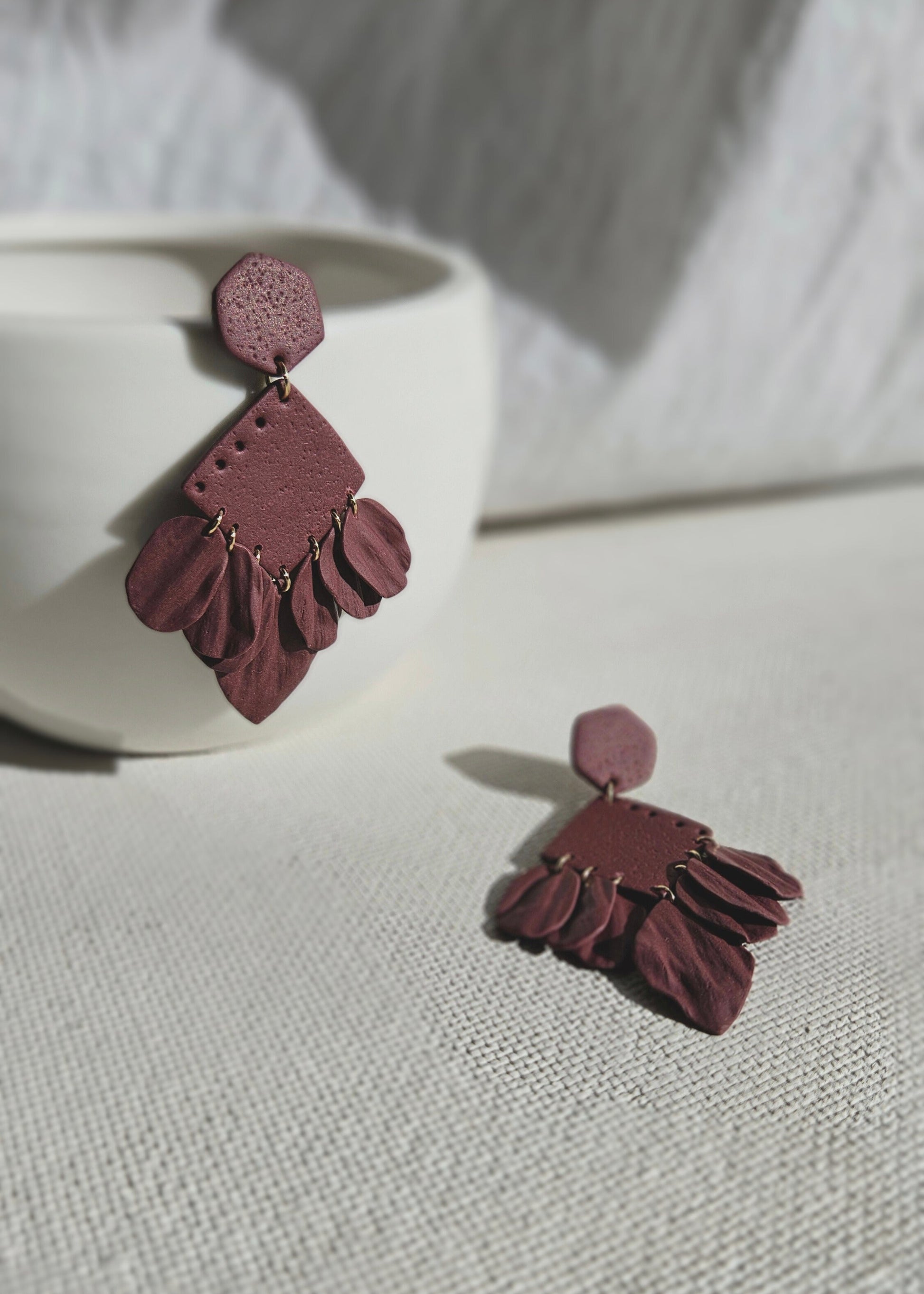La paire de boucles d'oreilles Epanouie dans son coloris Bordeaux est présentée à plat, devant un rideau