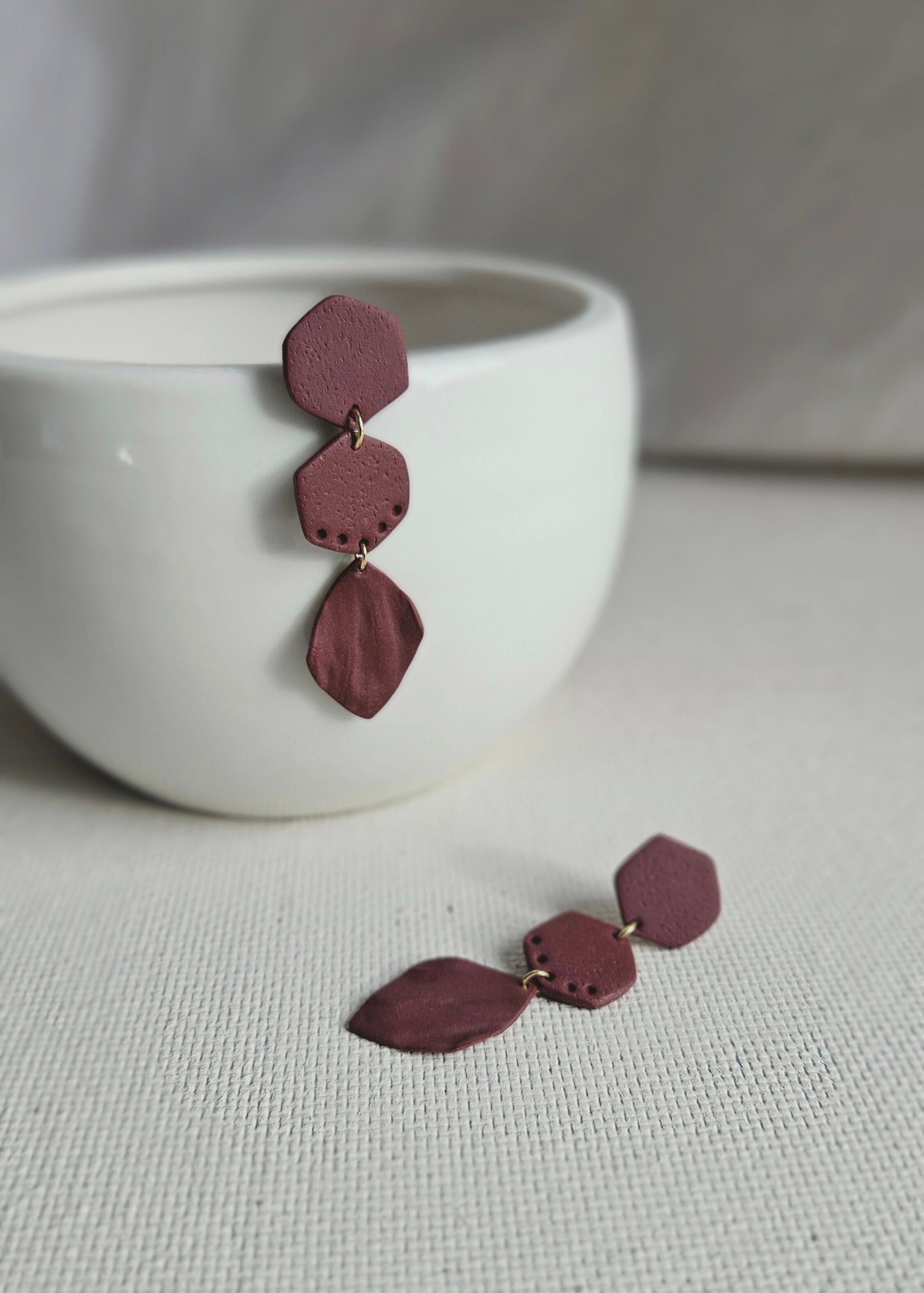 La paire de boucles d'oreilles Naturelle dans son coloris Bordeaux est présentée à plat, devant un rideau