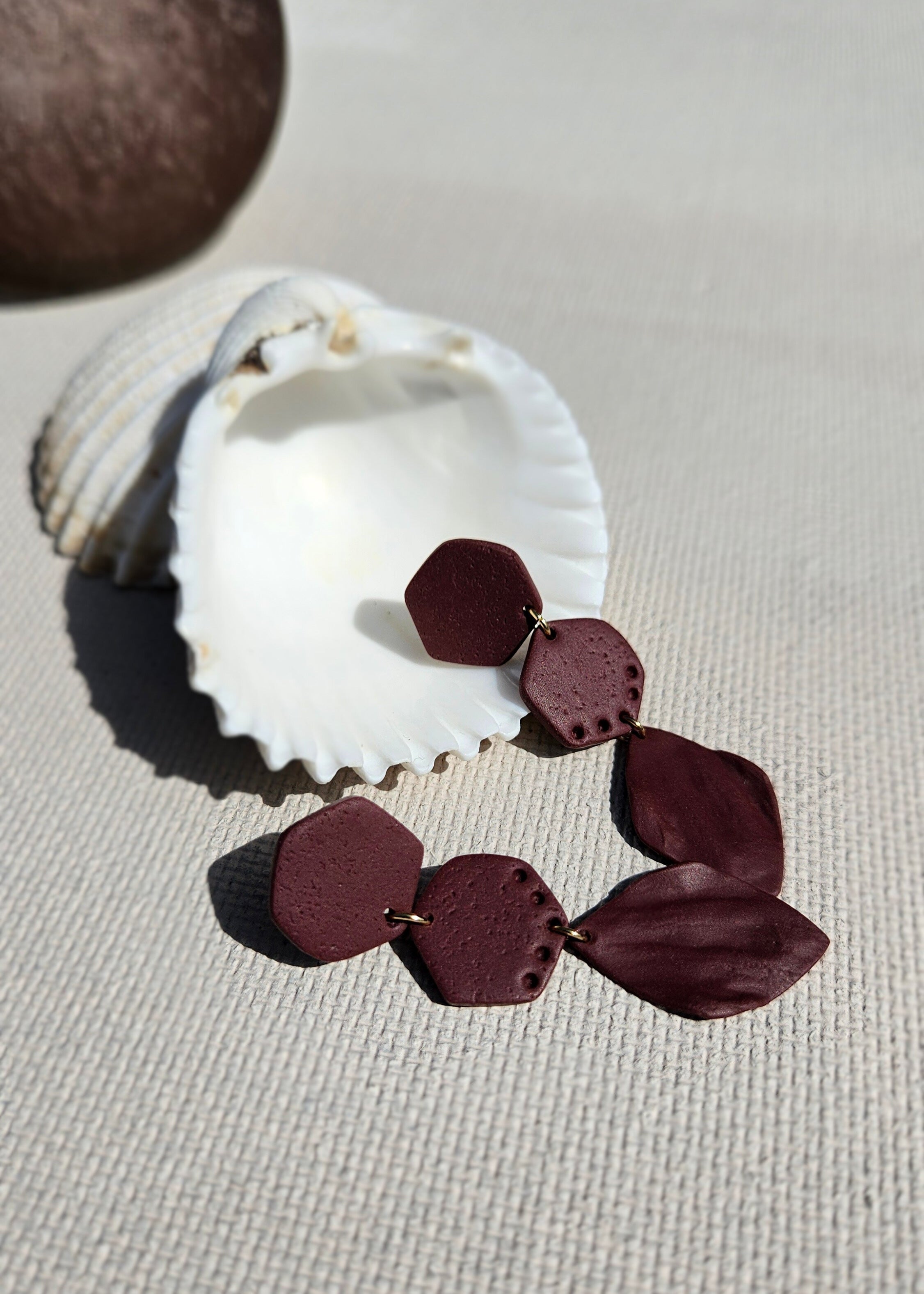 La paire de boucles d'oreilles Naturelle dans son coloris Bordeaux est présentée à plat, dans un coquillage
