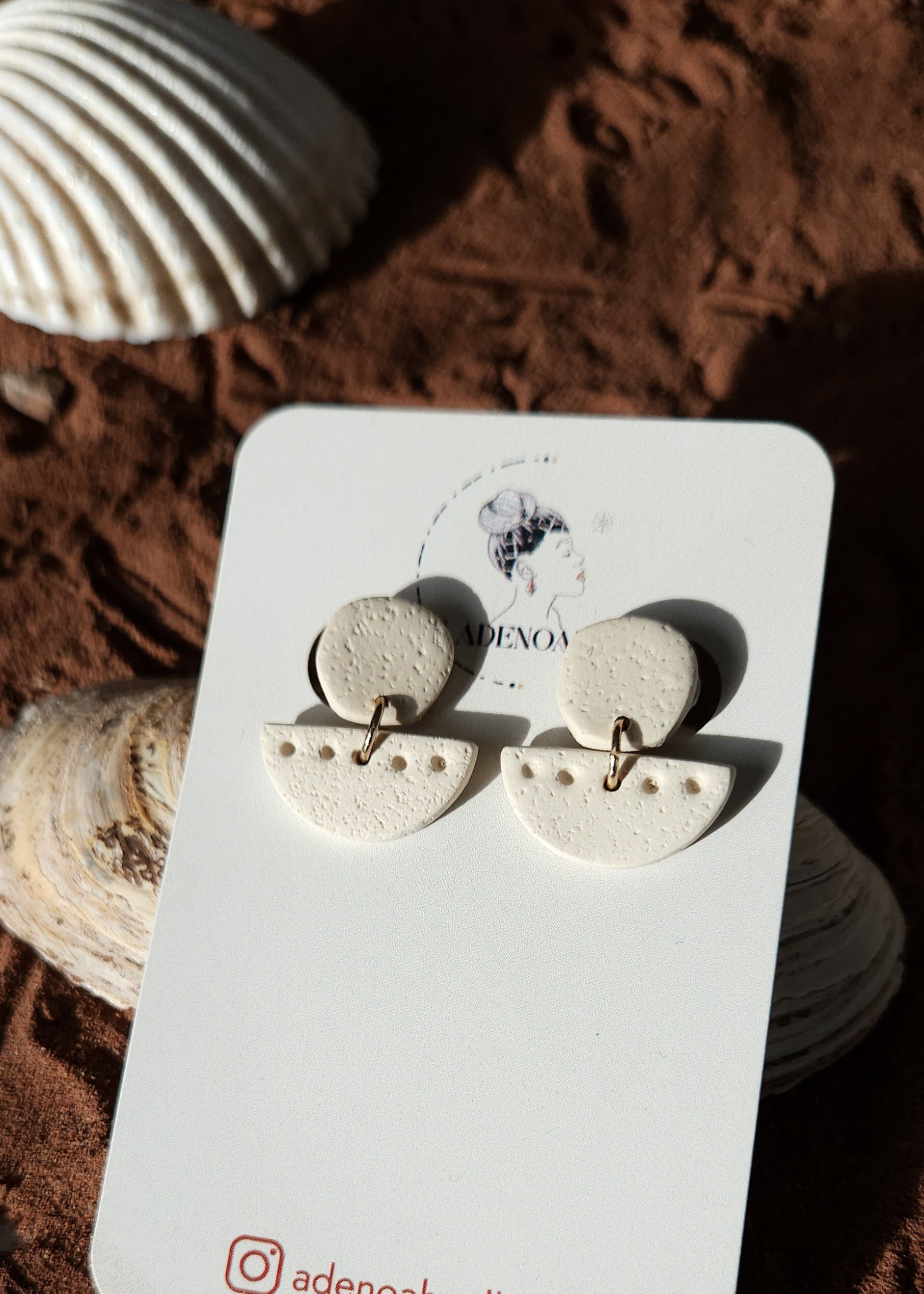 La paire de boucles d'oreilles Patiente dans son coloris sable est présentée à plat, dans le sable, devant un coquillage