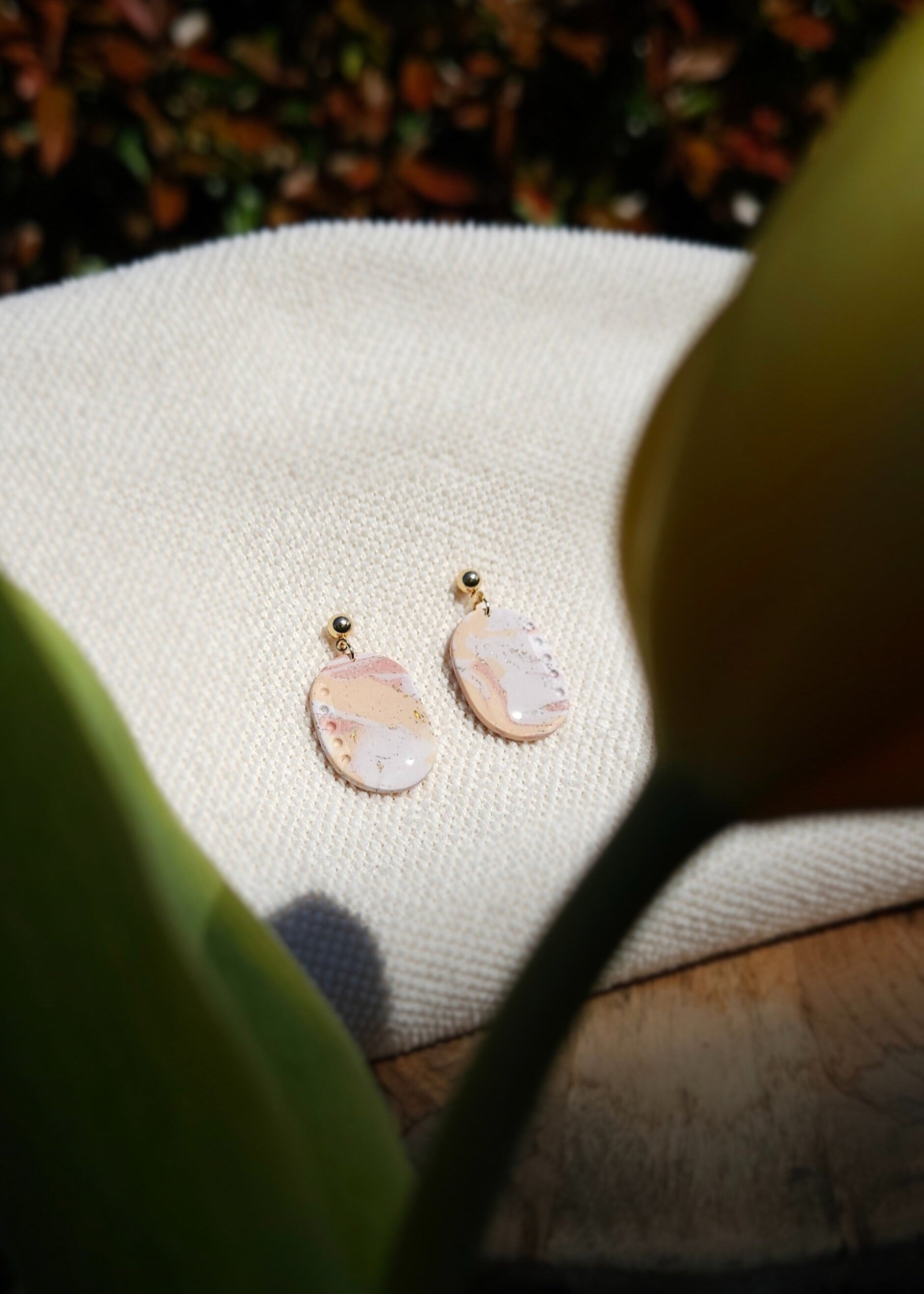 La paire de boucles d'oreilles Confiante dans son coloris Douceur poudrée mélangé à du Peau de pêche et du blanc est présentée à plat, sur un tabouret en bois accompagnée d'une tulipe jaune