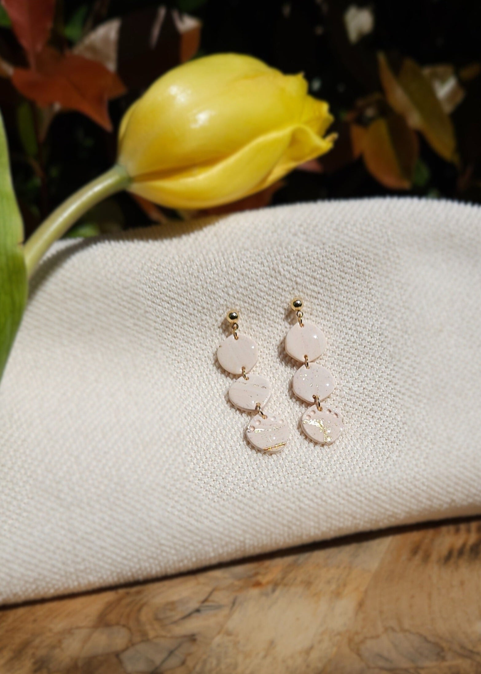 La paire de boucles d'oreilles Envoûtante dans son coloris Beige rosé est présentée à plat, sur un tabouret en bois accompagnée d'une tulipe jaune