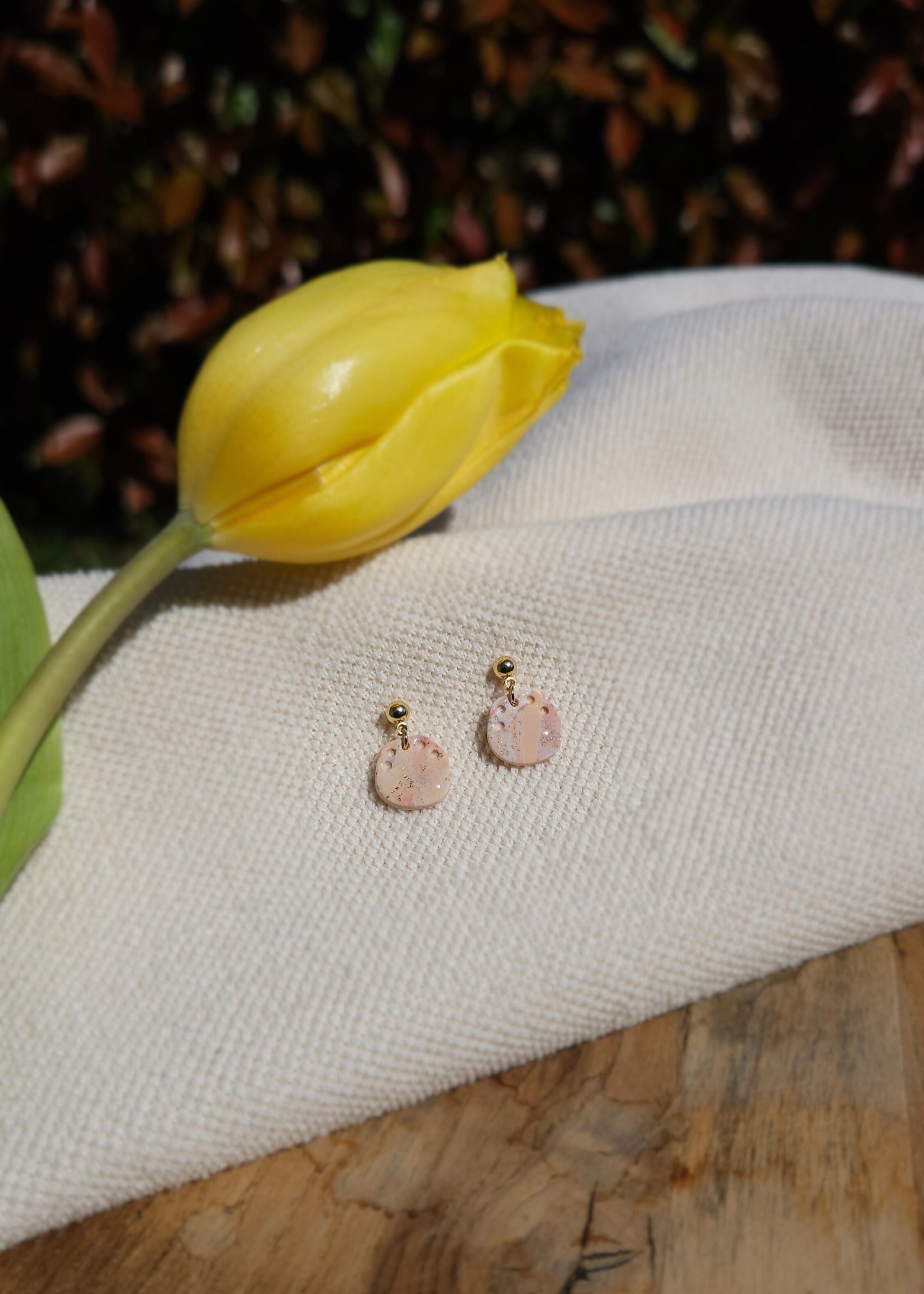 La paire de boucles d'oreilles Talentueuse dans son coloris Douceur poudrée mélangé à du Peau de pêche et du blanc est présentée à plat, sur un tabouret en bois accompagnée d'une tulipe jaune