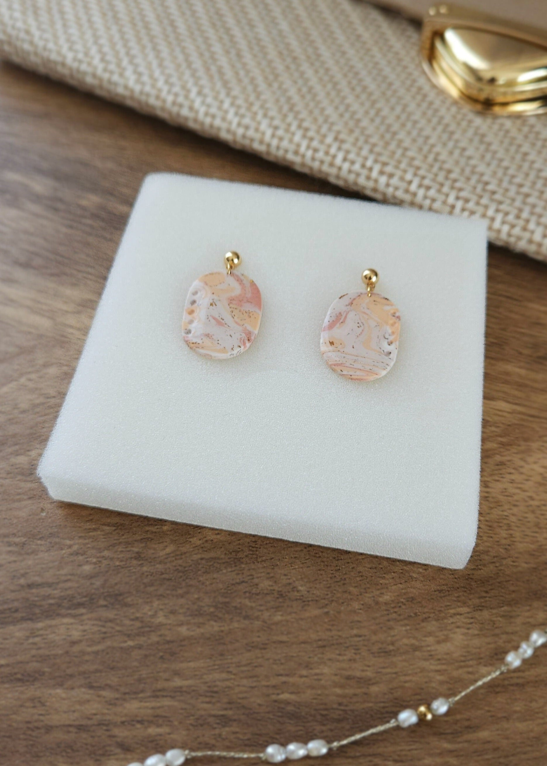La paire de boucles d'oreilles Confiante dans son coloris Douceur poudrée mélangé à du Peau de pêche et du blanc est présentée à plat, dans la mousse blanche de son écrin, à côté d'une pochette de fête