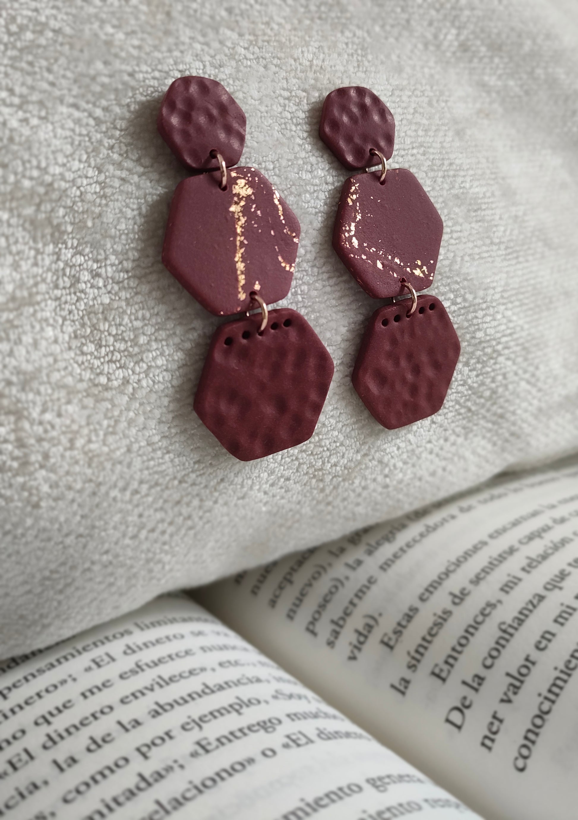 La paire de boucles d'oreilles Courageuse dans son coloris Bordeaux est présentée à plat, devant un feuillage aux tons verts et rouges