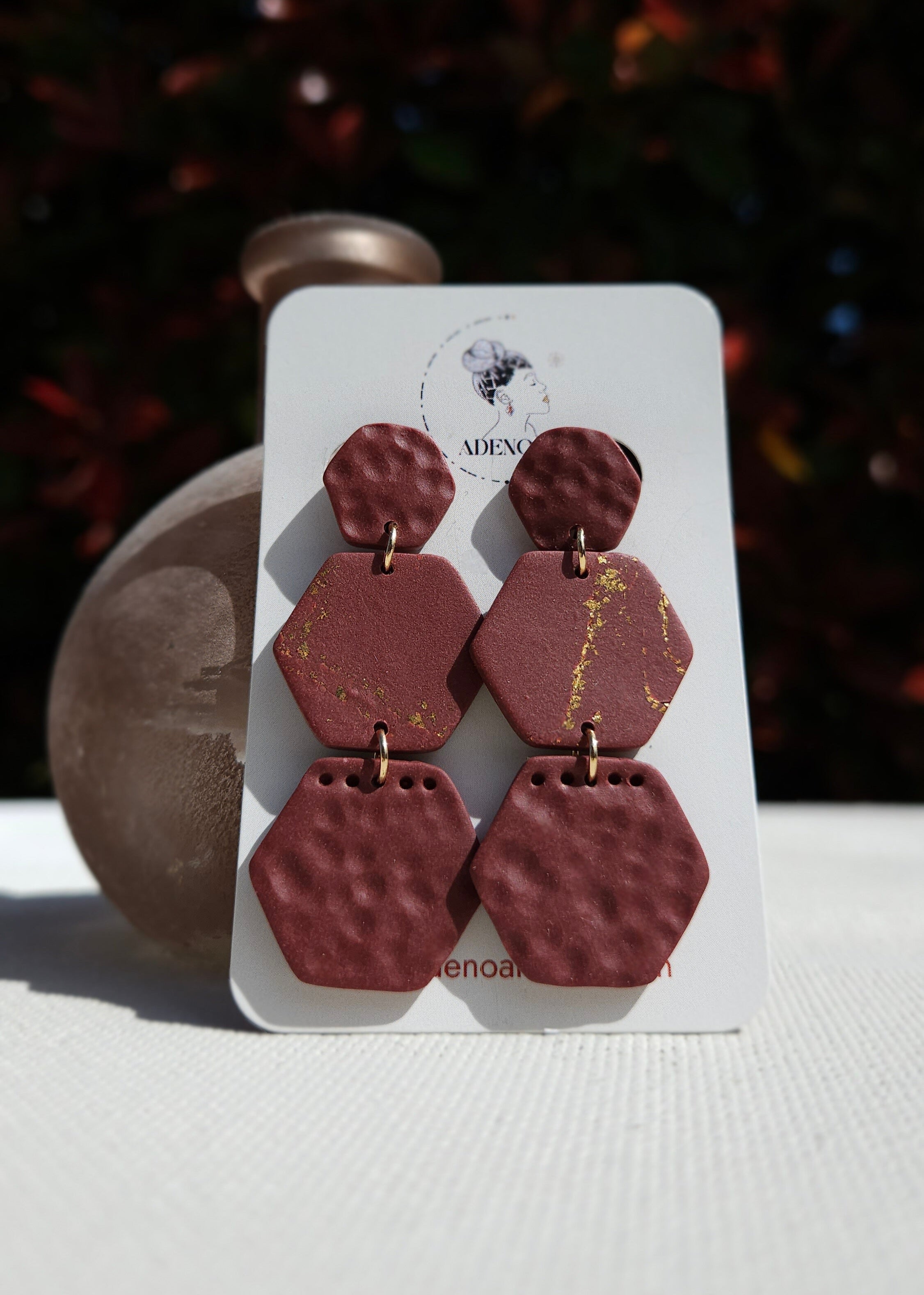La paire de boucles d'oreilles Courageuse dans son coloris Bordeaux est présentée à plat, devant un feuillage aux tons verts et rouges