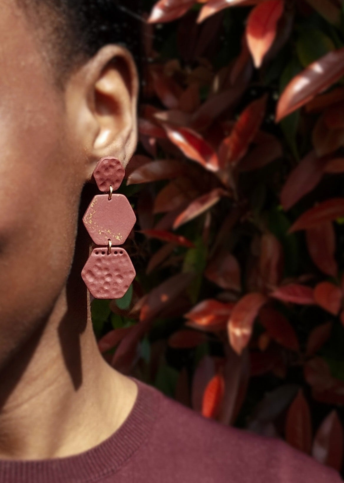 Le mannequin porte la paire de boucles d'oreilles Courageuse dans son coloris Bordeaux. Ce modèle est composé de trois parties externes bosselées et une pièce centrale lisse avec des éclats de feuille d'or