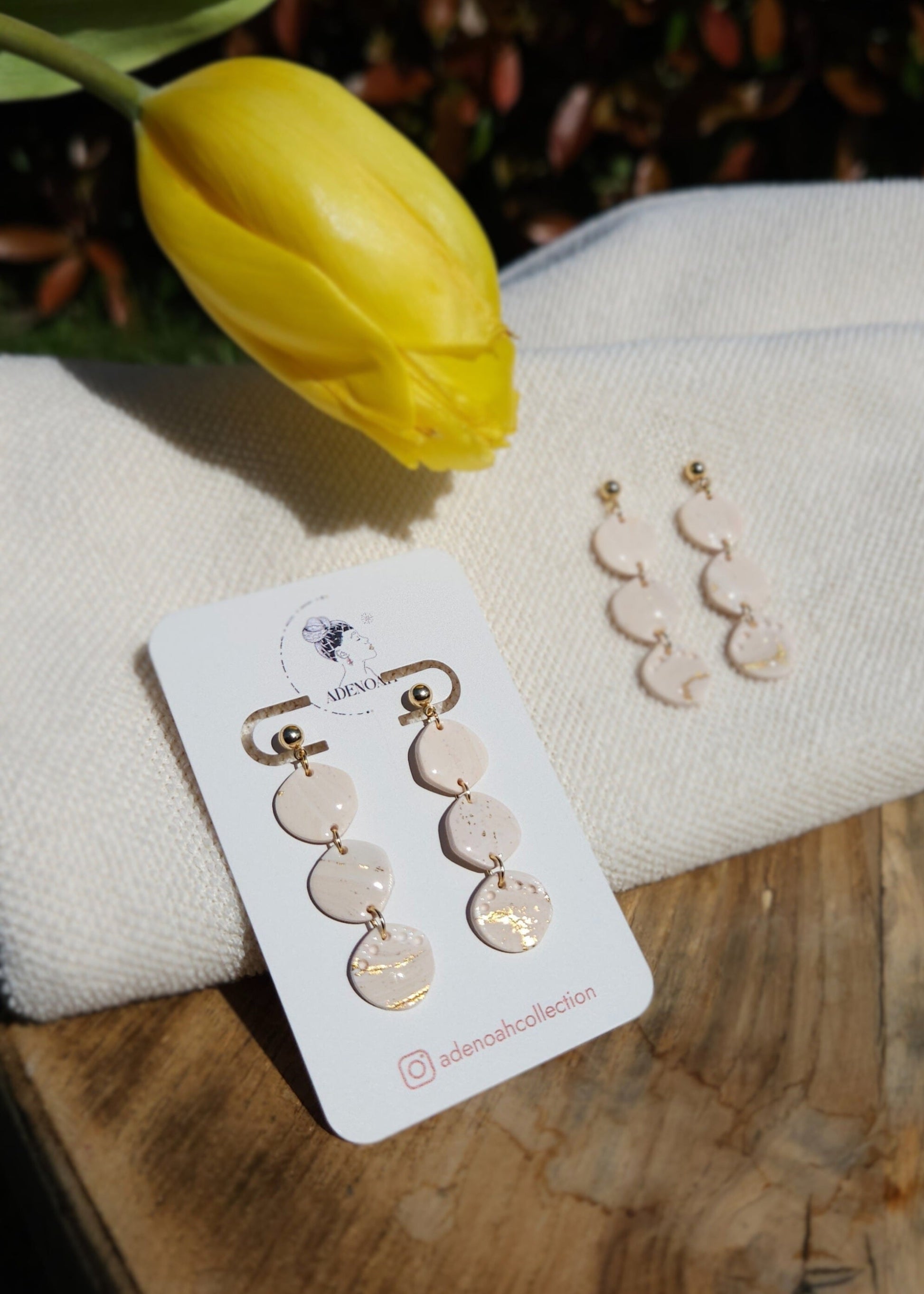 La paire de boucles d'oreilles Envoûtante dans son coloris Beige rosé est présentée à plat, sur un tabouret en bois accompagnée d'une tulipe jaune à côté d'un deuxième modèle sur cartonnette