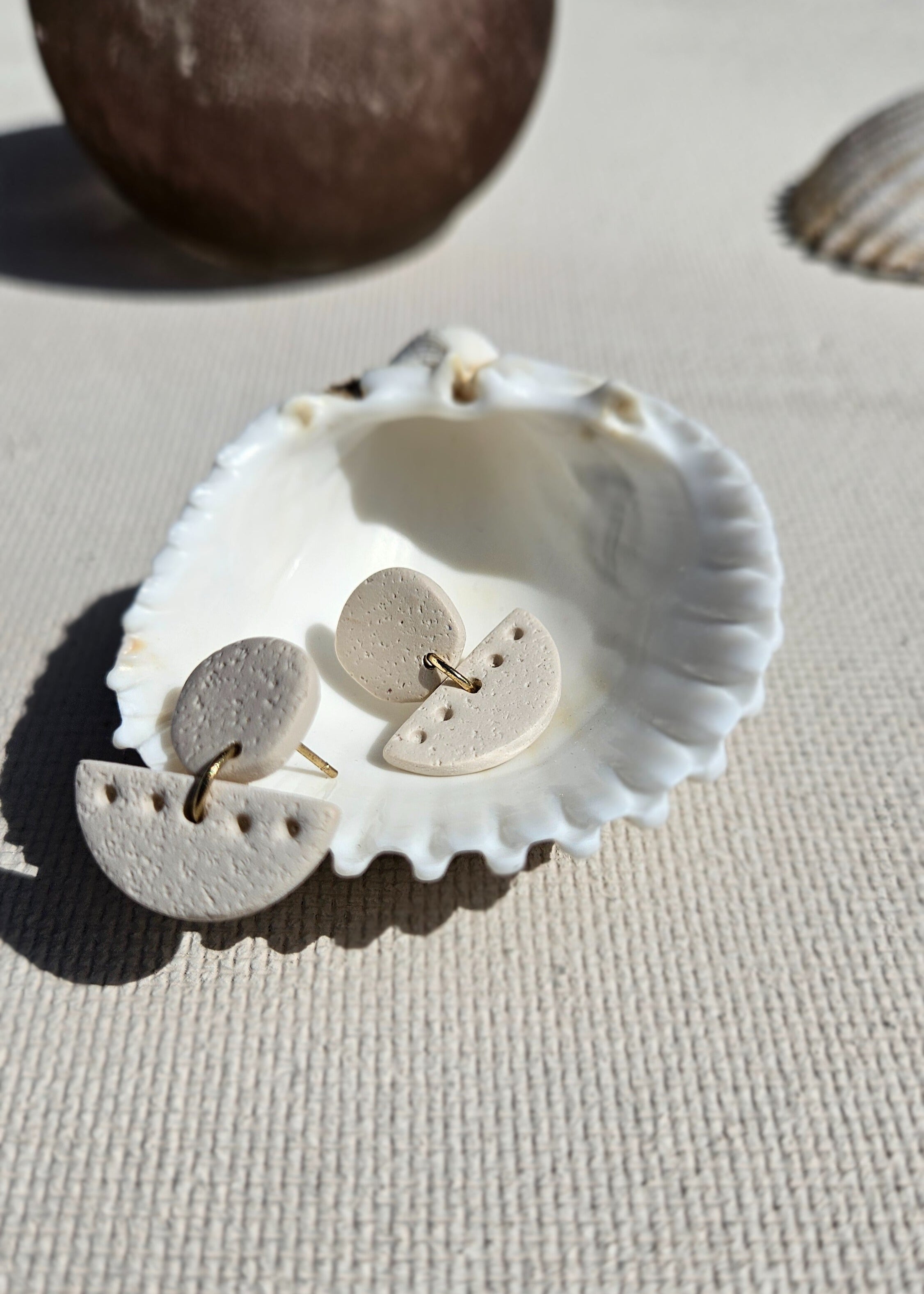 La paire de boucles d'oreilles Patiente dans son coloris sable est présentée à plat, dans un coquillage