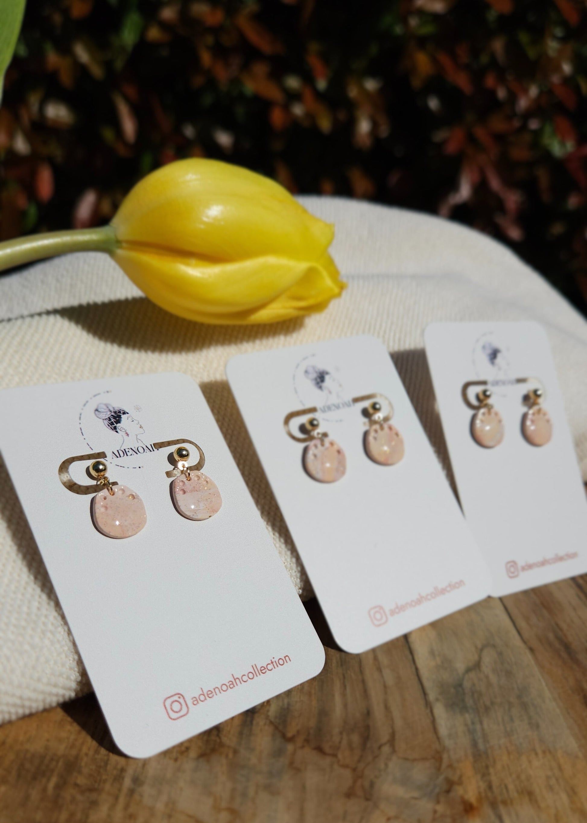 Trois paires de boucles d'oreilles Talentueuse dans leur coloris Douceur poudrée mélangé à du Peau de pêche et du blanc sont présentées sur cartonnette, sur un tabouret en bois accompagnée d'une tulipe jaune